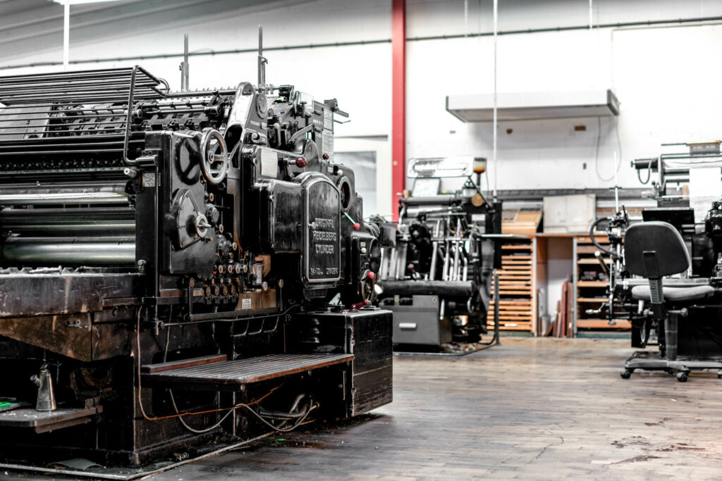 Schwarze, klassische Druckmaschinen der Firma Heidelberg in den Hallen der Druckerei Hartmann in Hilzingen. Dunkler Holzboden und weiße Wände im Hintergrund.